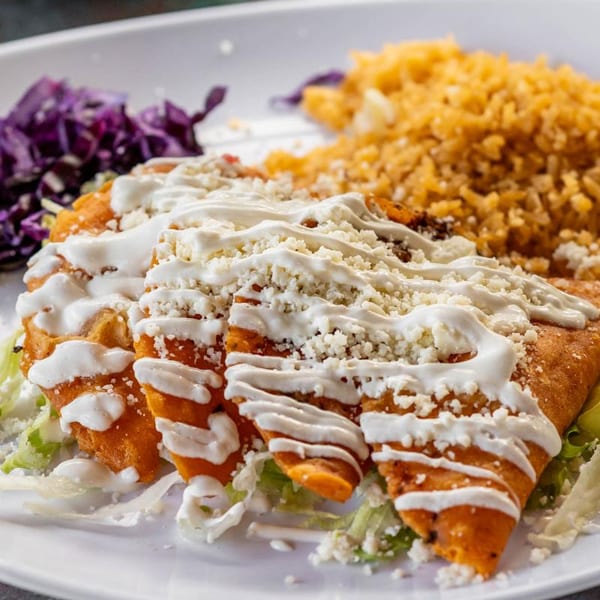 Three golden-fried enchiladas topped with sour cream and crumbled cheese, served with rice and purple cabbage.