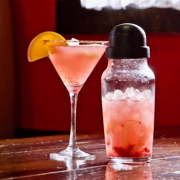Pink martini with orange slice and a cocktail shaker with ice and pink drink on a wooden bar.