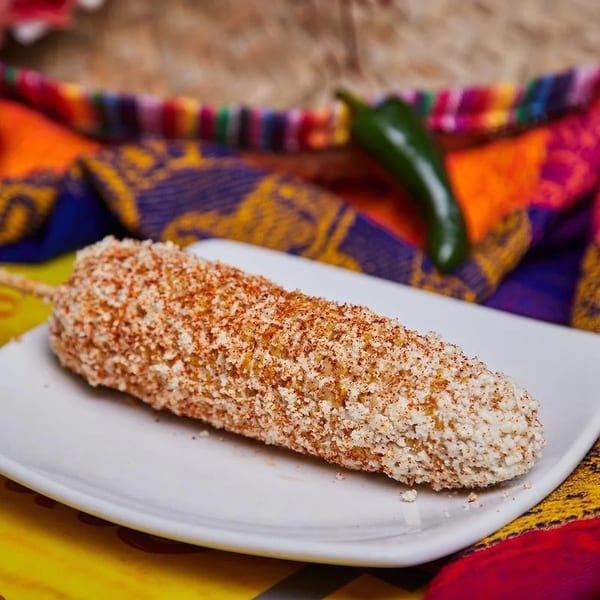 Elote, Mexican street corn, on a white plate with a colorful textile and jalapeño in the background.