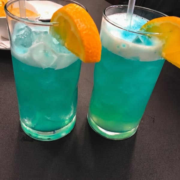 Two vibrant blue cocktails with orange slices and straws sit on a dark table.