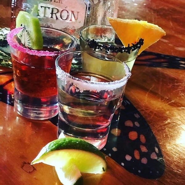 Three colorful Patron tequila shots with fruit garnishes and a lime wedge on a wooden table.