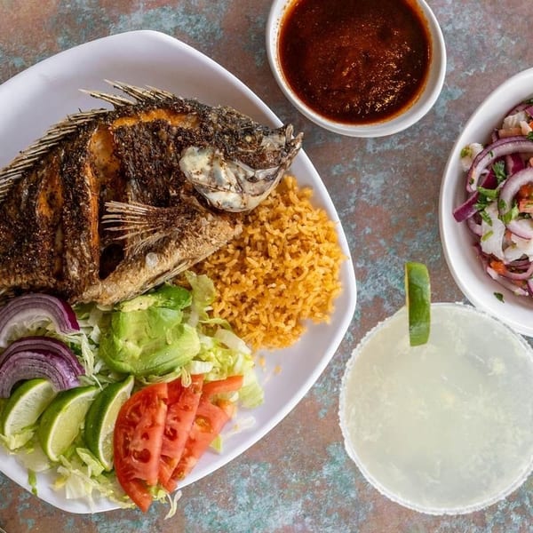 Fried whole fish with rice, salad, salsa, ceviche, and a margarita on a textured surface.
