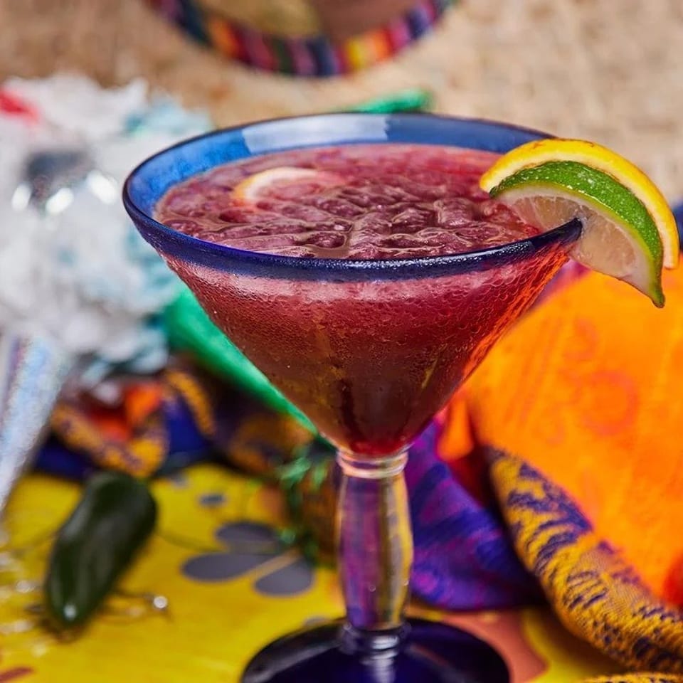A vibrant red frozen margarita in a blue-rimmed glass, garnished with lime and lemon slices.