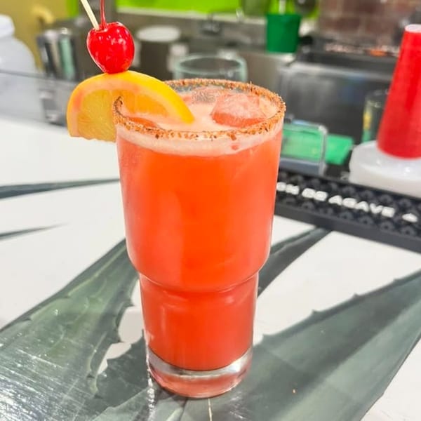 A vibrant red cocktail with a salted rim, garnished with an orange slice and cherry, on a bar.