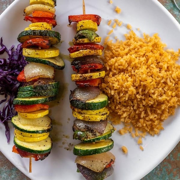 Two grilled vegetable skewers with zucchini, squash, peppers, and onions, served with yellow rice.