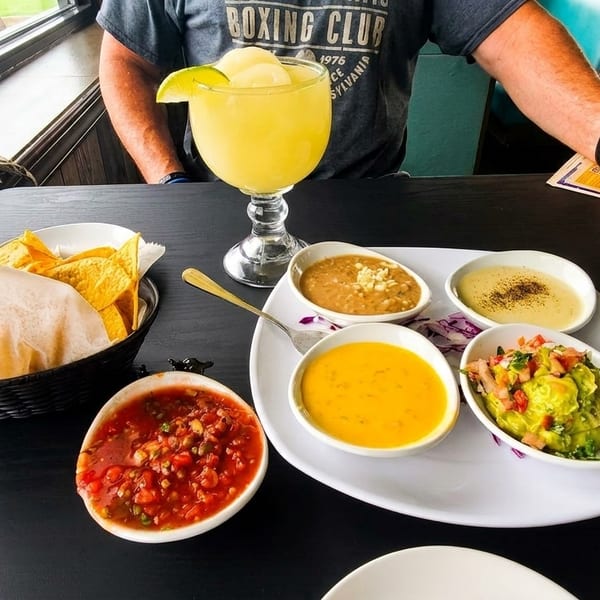 A person's arms are visible at a table with chips, salsa, guacamole, and other dips.