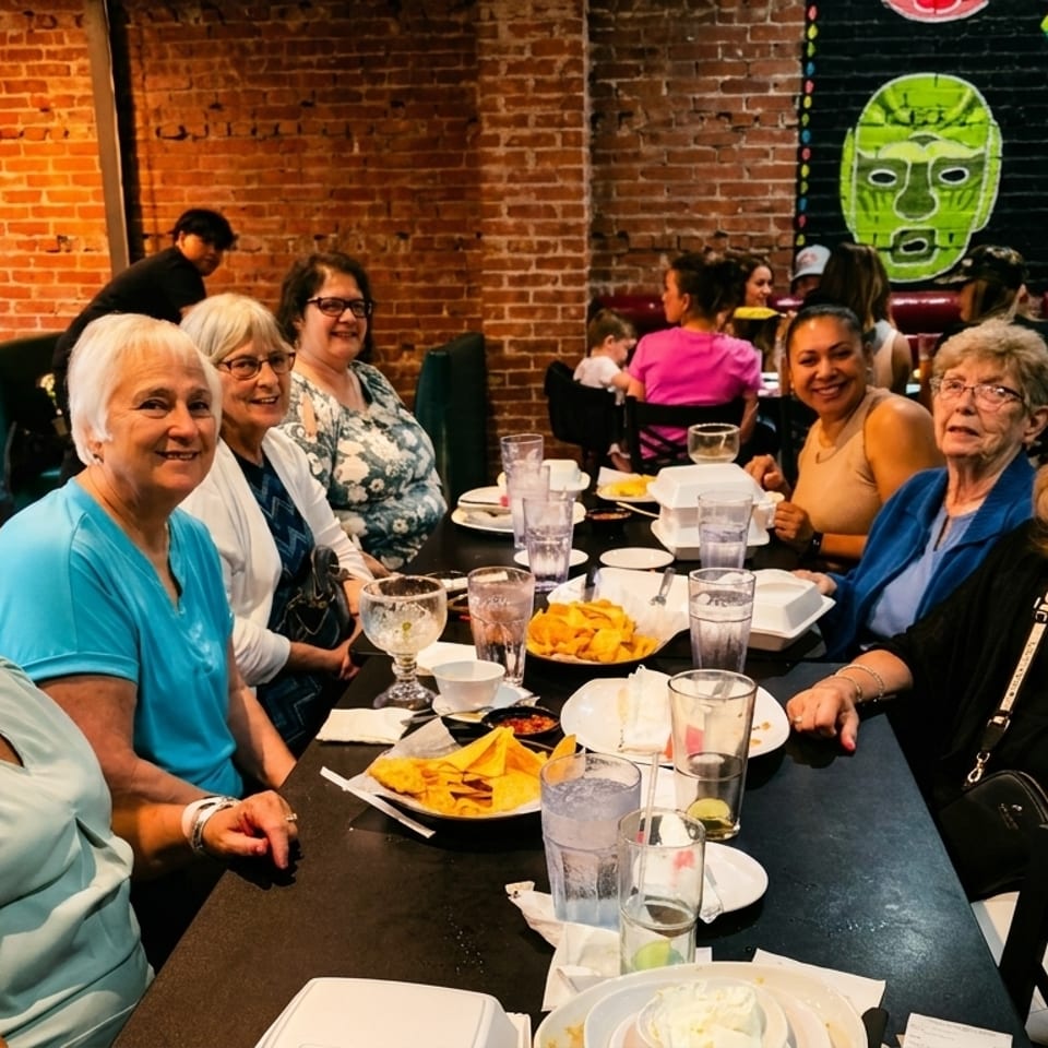 A group of smiling women enjoying a meal together at a restaurant with brick walls and colorful decor.